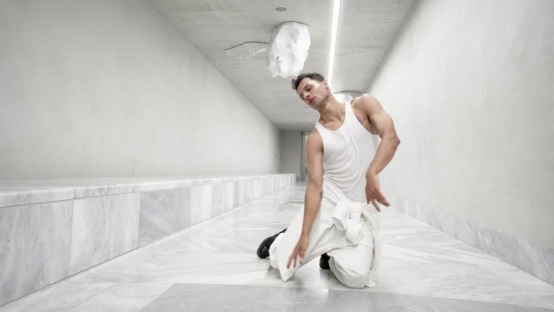 Adult male in white tank top and trousers kneeling in modern corridor with white walls and marble floor; sculpture on ceiling.