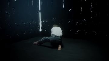 Adult male in a white shirt and blue jeans is kneeling on a dark floor in a dim indoor space, surrounded by metal hooks.