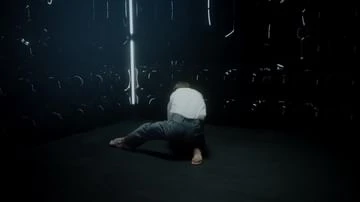 Adult male in a white shirt and blue jeans is kneeling on a dark floor in a dim indoor space, surrounded by metal hooks.