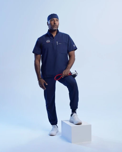 Full shot of an adult male wearing a navy blue scrub suit and sneakers, posing in a photo studio with a stethoscope and a white box.