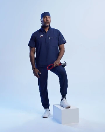 Full shot of an adult male wearing a navy blue scrub suit and sneakers, posing in a photo studio with a stethoscope and a white box.