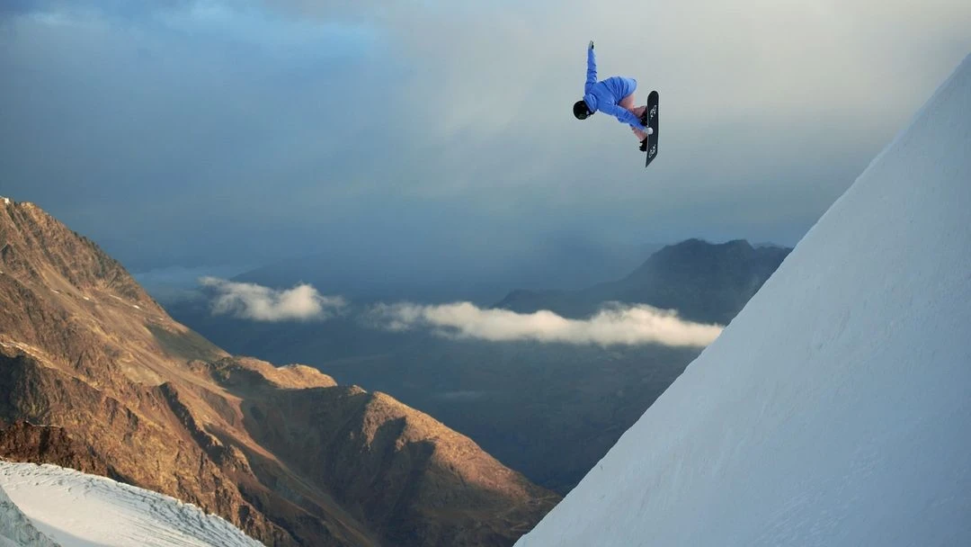 Snowboarder performs aerial stunt on snowy mountain peak with cloudy sky in background. Extreme winter sport action in alpine landscape.
