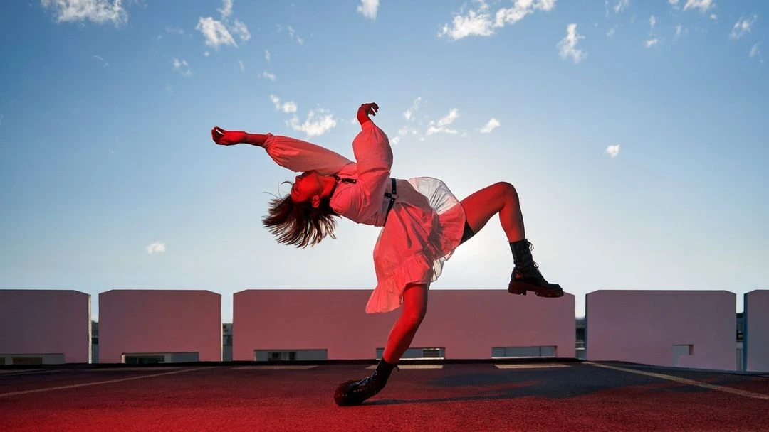 Young woman in mid-air doing an acrobatic pose on a red surface with white architectural shapes and a cloudy blue sky in the background.