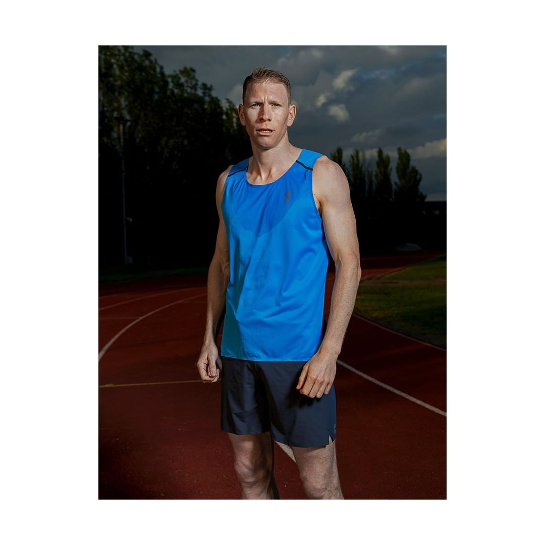Male athlete stands on a running track in a bright blue tank top and navy running shorts on a cloudy day, captured in a medium shot.