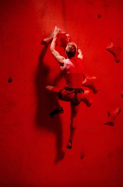 A man climbs an indoor, red climbing wall, using safety gear. He is viewed from the back, with shadow on the textured wall.