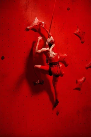 An adult female rock climber ascends a red indoor climbing wall with handholds and a safety rope. The athlete is dressed in red activewear.