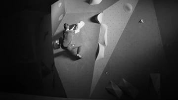 Monochrome shot of a person bouldering indoors on a geometric climbing wall, captured in a dimly lit, grayscale setting.
