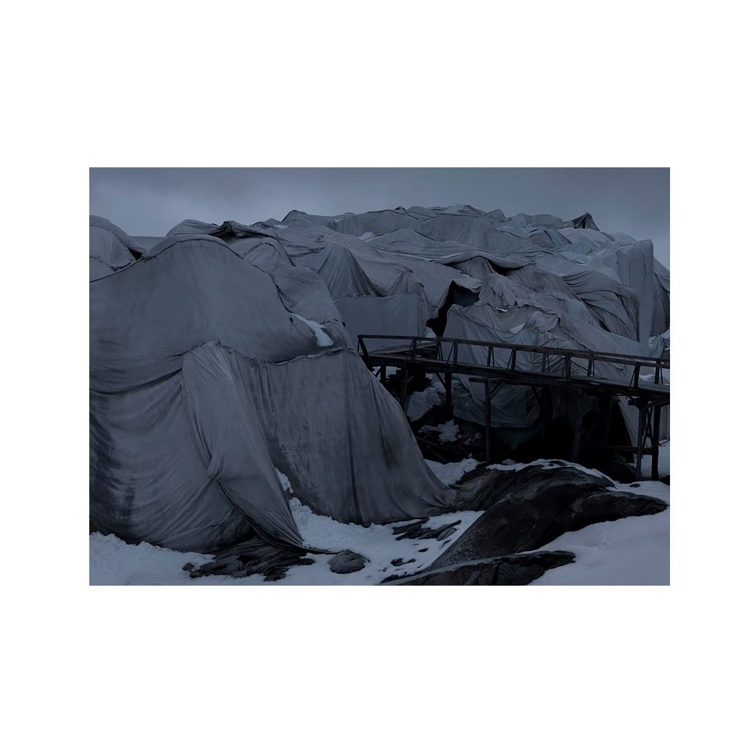 A landscape featuring gray rocks and snow covered with gray tarps and a wooden bridge, under an overcast sky, creating a somber scene.