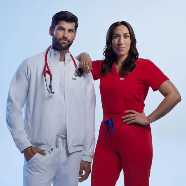 Two healthcare workers in scrubs: a man in a white jacket with a stethoscope, and a woman in red scrubs, stand in a bright studio setting.
