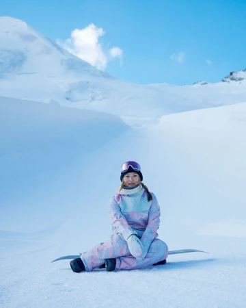 Female snowboarder sits on snowy slope wearing goggles and pastel snowsuit, with snow-capped mountain range against a blue sky.