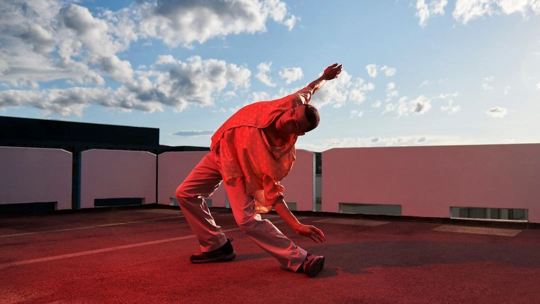 Man in red clothing performs a contemporary dance pose on a rooftop under a blue sky with scattered clouds during the daytime.