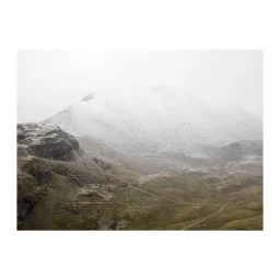Misty mountain landscape with snow-dusted rocky terrain and a winding dirt path under an overcast sky.