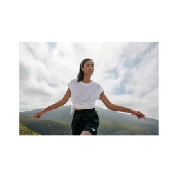 Woman wearing white t shirt and black shorts posing outdoors against a mountainous landscape and cloudy sky.