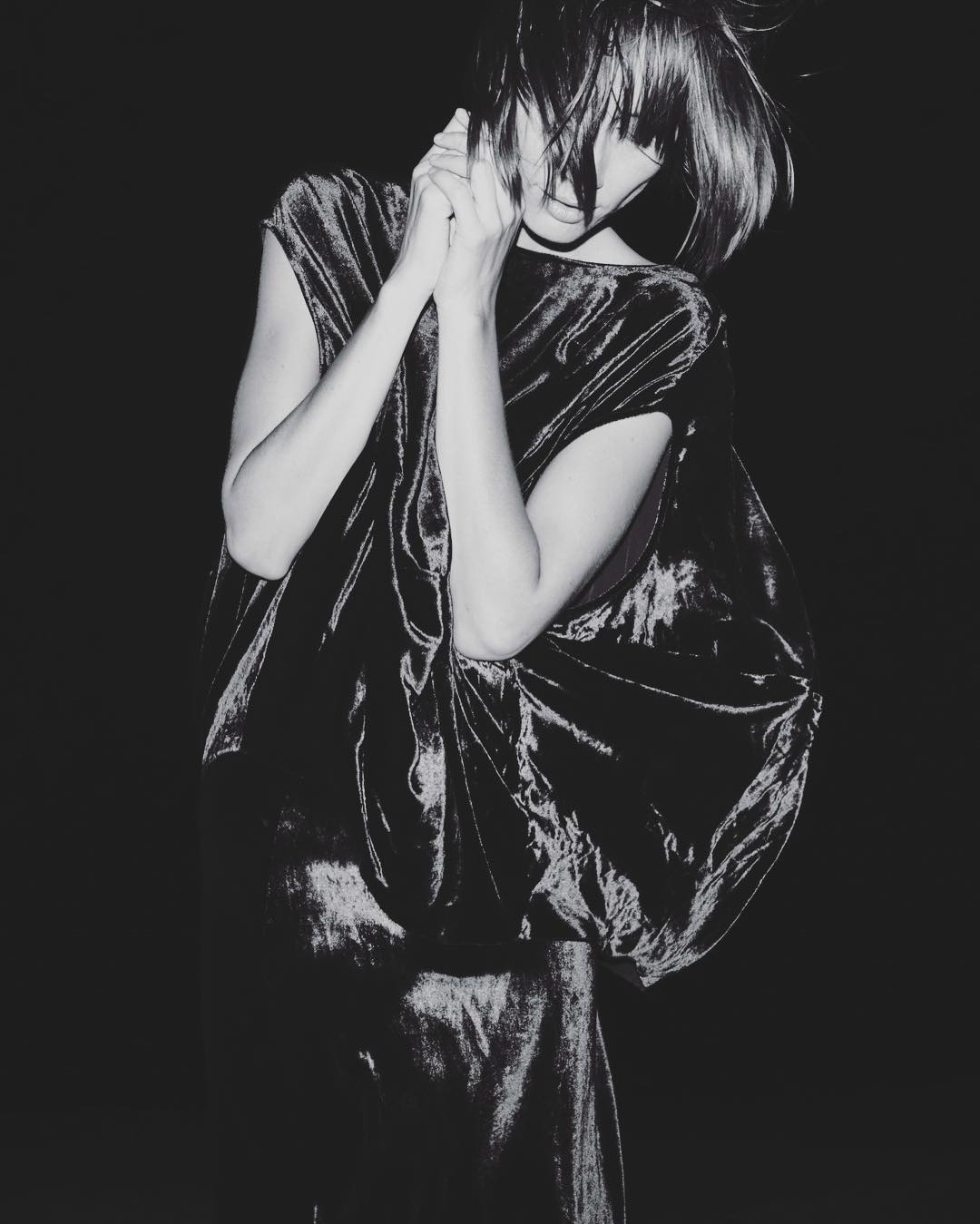 Monochrome portrait of a woman with short dark hair wearing a shiny black dress in a dark indoor setting, her hands near her face.