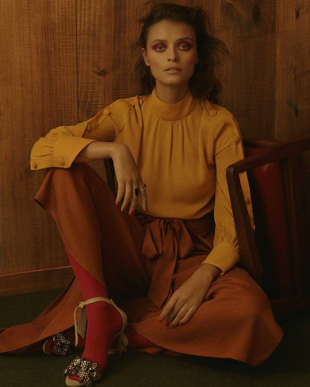 Fashionable woman poses indoors wearing a yellow blouse, high-waisted rust-colored pants, red socks, and embellished heeled shoes.