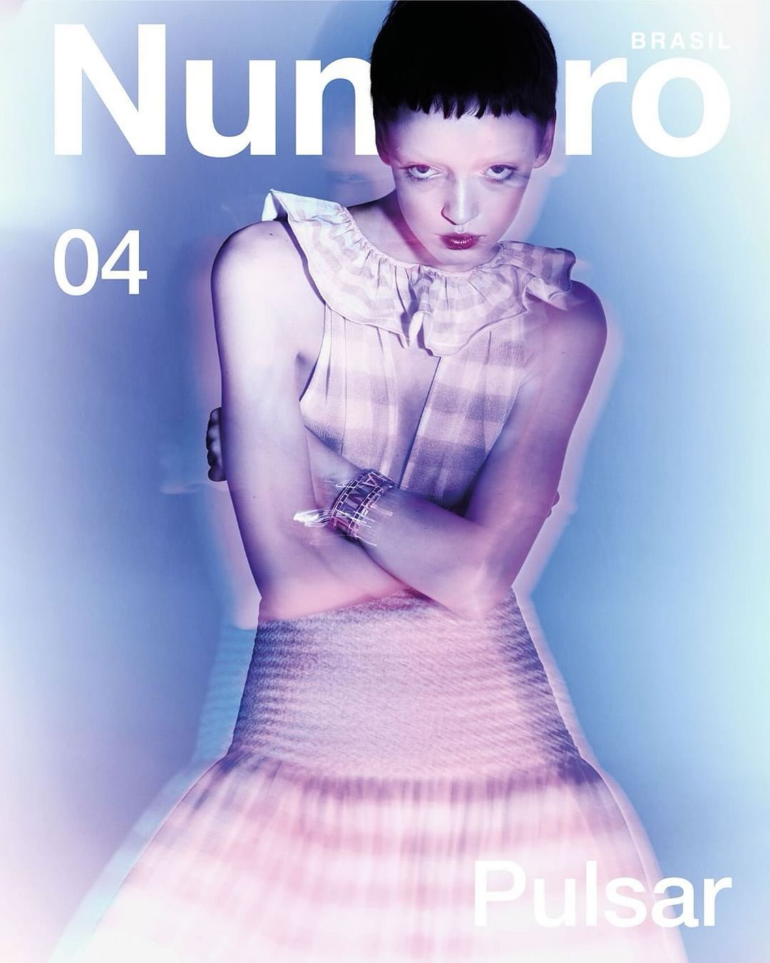 Fashion magazine cover featuring a woman with crossed arms wearing a dress with a ruffled collar, set against a blurred light blue background.