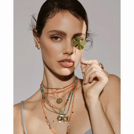 Close-up of a woman with multiple necklaces, gold jewelry, and a four-leaf clover covering her eye against a neutral backdrop.