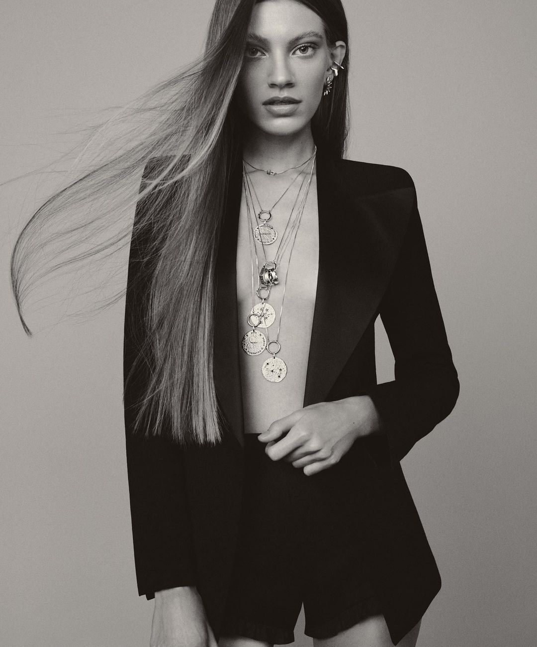 Monochrome studio shot of a woman in a black jacket and shorts, adorned with multiple layered necklaces against a grey backdrop.