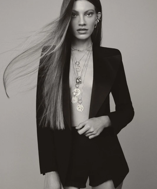 Monochrome studio shot of a woman in a black jacket and shorts, adorned with multiple layered necklaces against a grey backdrop.