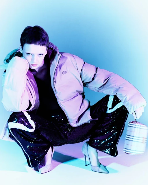 Fashion shot of a woman with short dark hair wearing a puffer jacket and sequined pants squatting with a bucket bag in a studio setting.