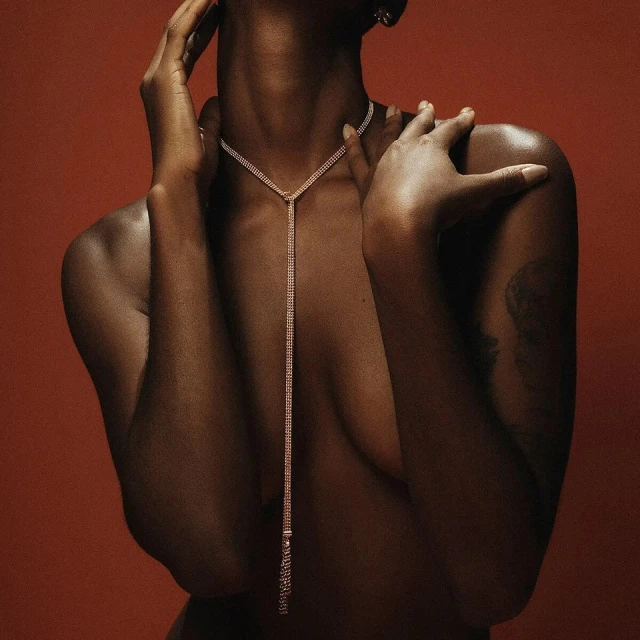 Portrait of a Black woman with bare shoulders, wearing a long, shimmering Y-shaped necklace against a dark red background.