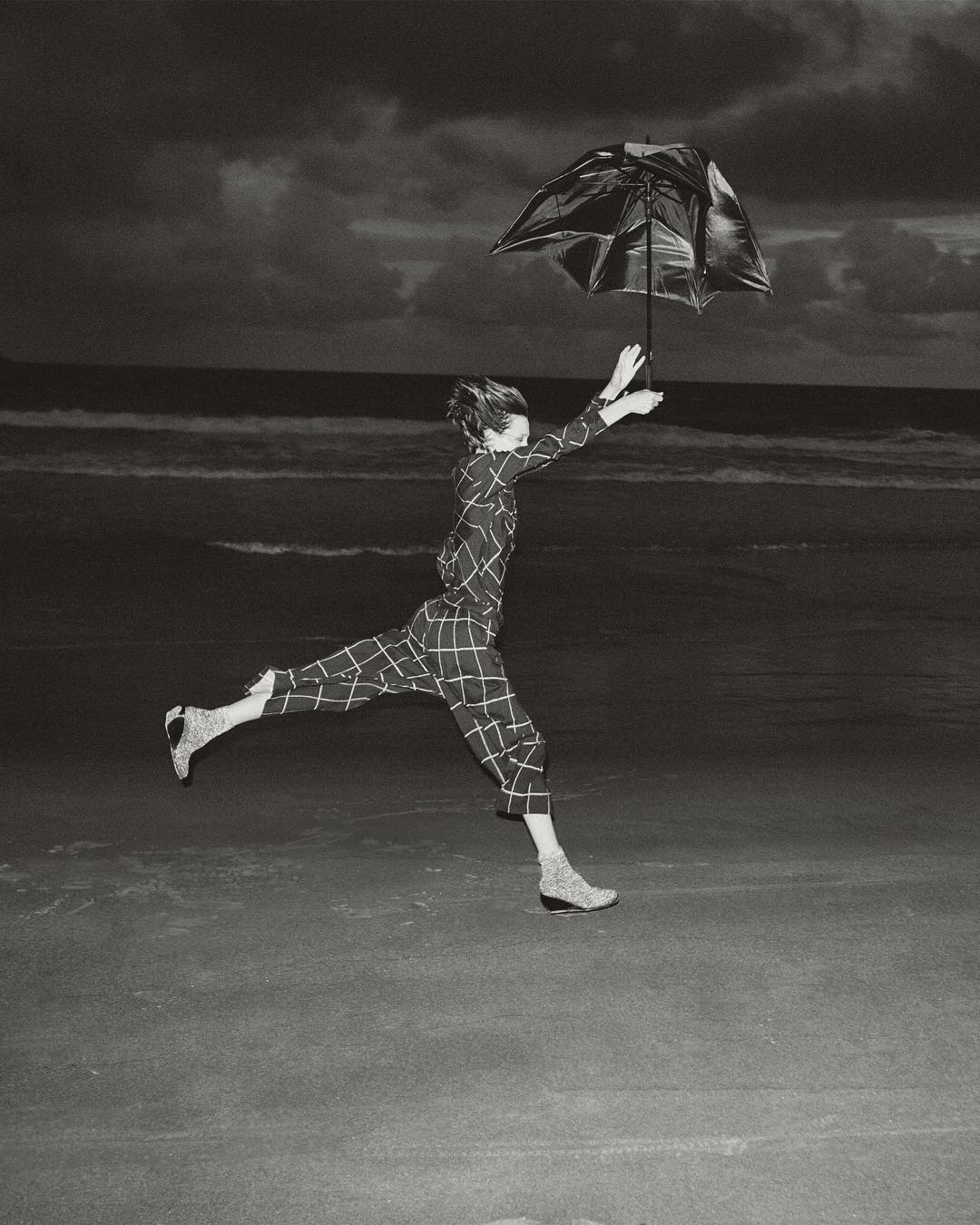 Monochrome shot of an adult running on a beach holding an umbrella against a dark sky with waves. Windblown fashion and movement.