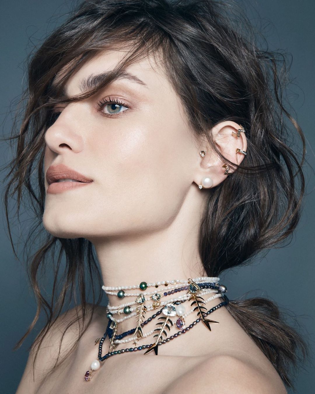 Profile shot of a brunette woman with pearl jewelry including layered pearl choker necklaces and multiple pearl earrings, grey studio backdrop.