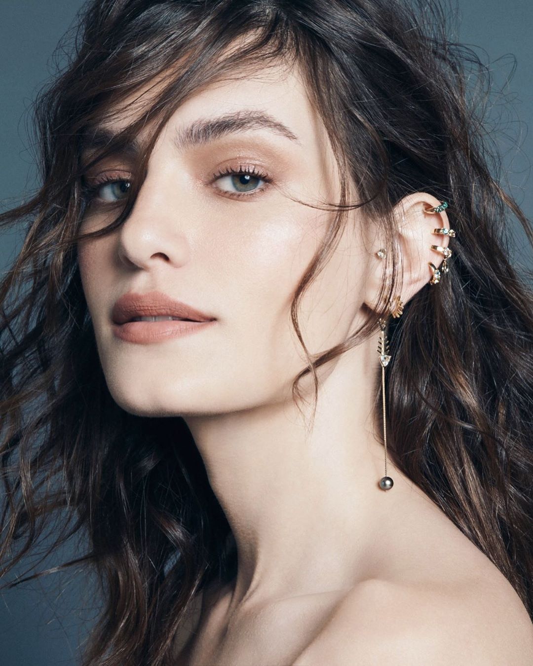 Portrait of a woman with long brunette hair wearing an ornate ear cuff and dangling earrings against a grey background in soft light.
