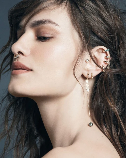 Close-up of an adult woman with a nose ring and multiple ear piercings with gold jewelry against a soft grey background in a studio.