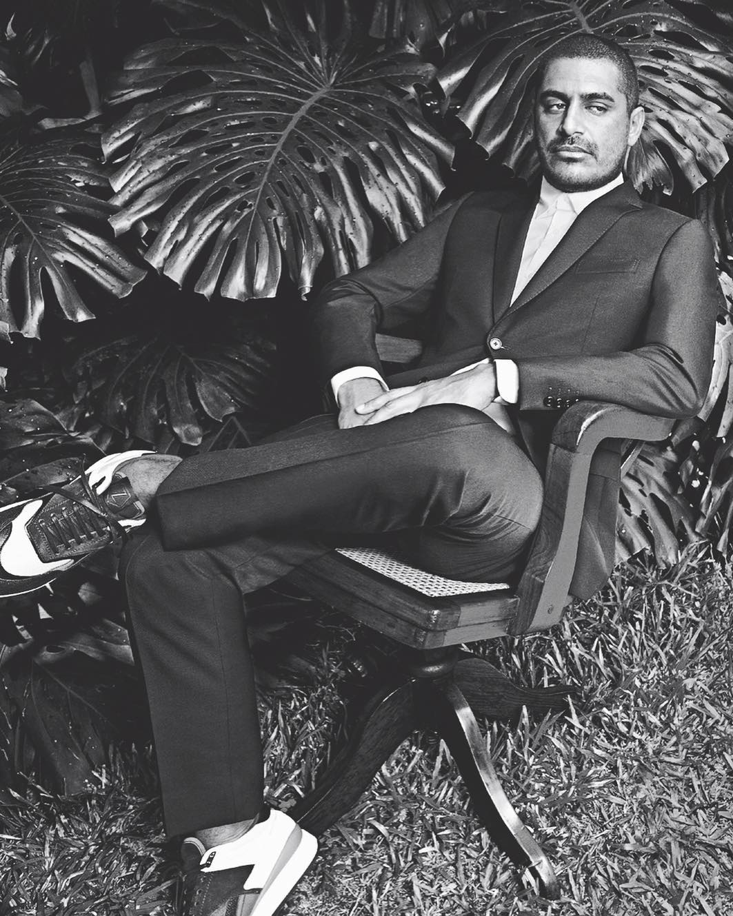 Black and white portrait of a man in a suit sitting in a chair outdoors against a leafy backdrop, with sneakers, facing camera.