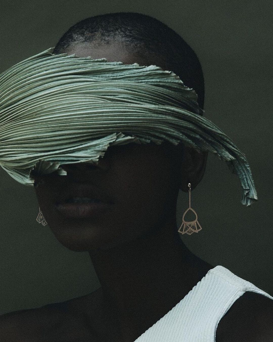 Elegant studio portrait of a bald African-American female model with statement earrings, face partially covered by a green pleated textile.