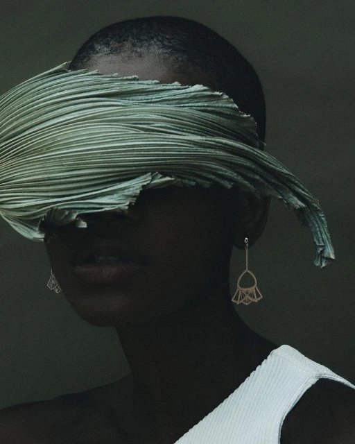 Elegant studio portrait of a bald African-American female model with statement earrings, face partially covered by a green pleated textile.