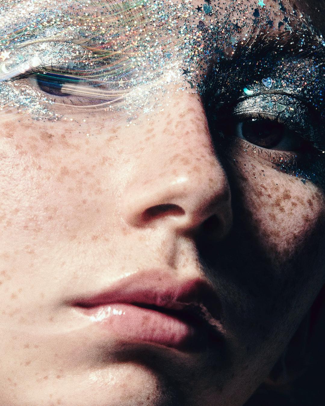 Close-up beauty shot of an adult female's face with glitter eye makeup, freckles, and neutral lips against a blurred background.