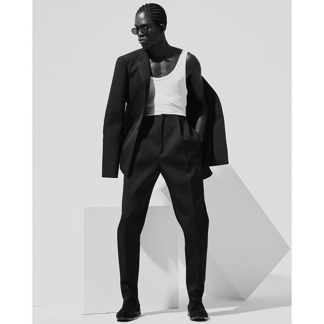 Studio shot of an adult male model in black suit, white top, and sunglasses posing against a white geometric structure.