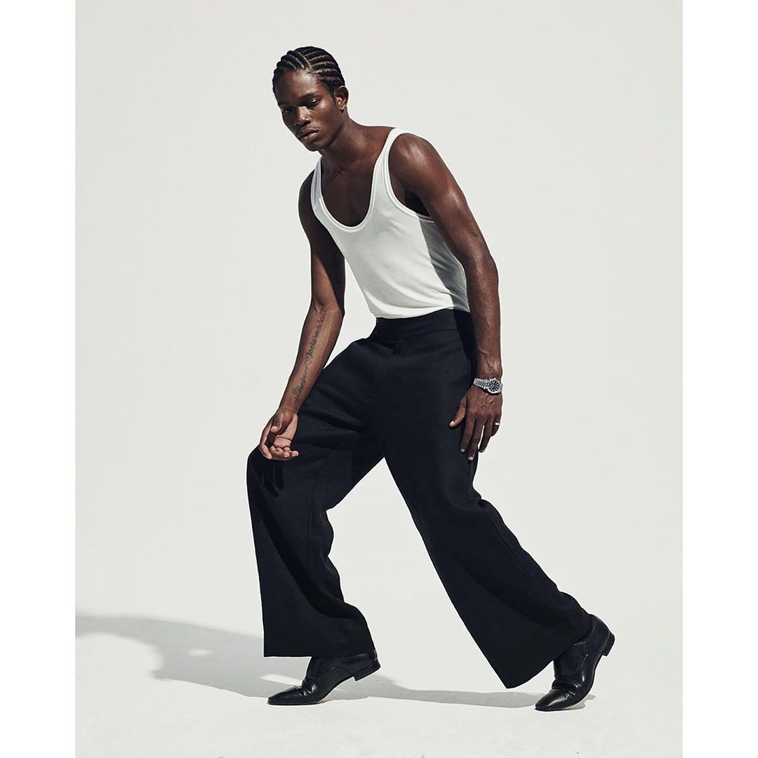 An athletic Black male fashion model poses in a white tank top and black dress pants on a white background in a studio shot.