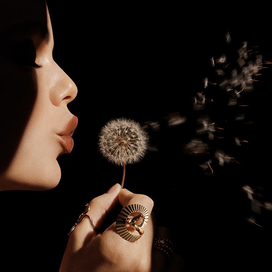 Woman blowing on dandelion seeds in dark studio. Close-up of model's face, hand with rings, dandelion seeds scattering, dark background.
