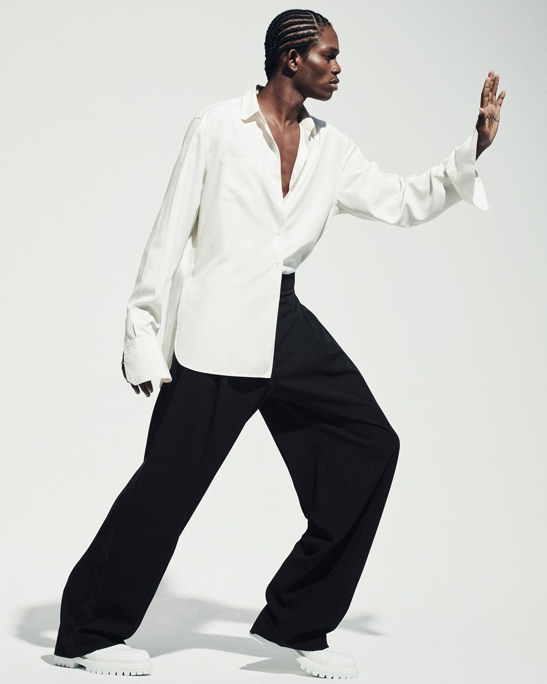 Fashion shot of a Black male model with cornrows, wearing a white shirt and black trousers, standing in a dynamic pose against a white background.