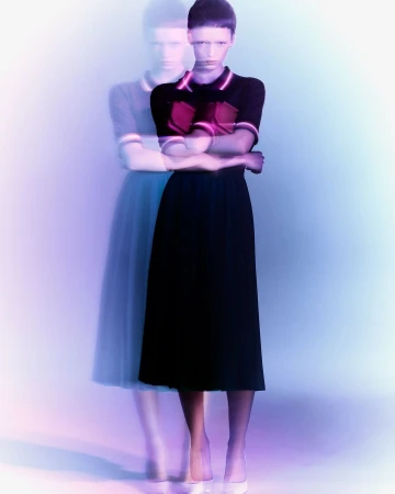 A woman stands with her arms crossed in a short-sleeved top and long skirt, with an out-of-focus duplicate of herself behind her.