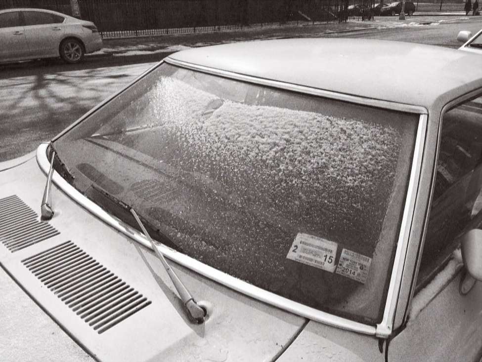 A vintage car's windshield covered in snow with its wipers up, parked on a city street with a car and trees in the background, black and white.