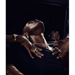 Close-up portrait of a person with head tilted back, wearing gemstone rings, gold bracelet, earrings, and a delicate necklace against a dark ground.