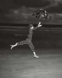 Model in checkered trousers leaping on a sandy beach at night while holding an open umbrella, photographed by Gil Inoue.