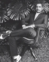 Portrait of a man in a dark suit sitting on a wooden chair outdoors surrounded by large tropical foliage leaves.