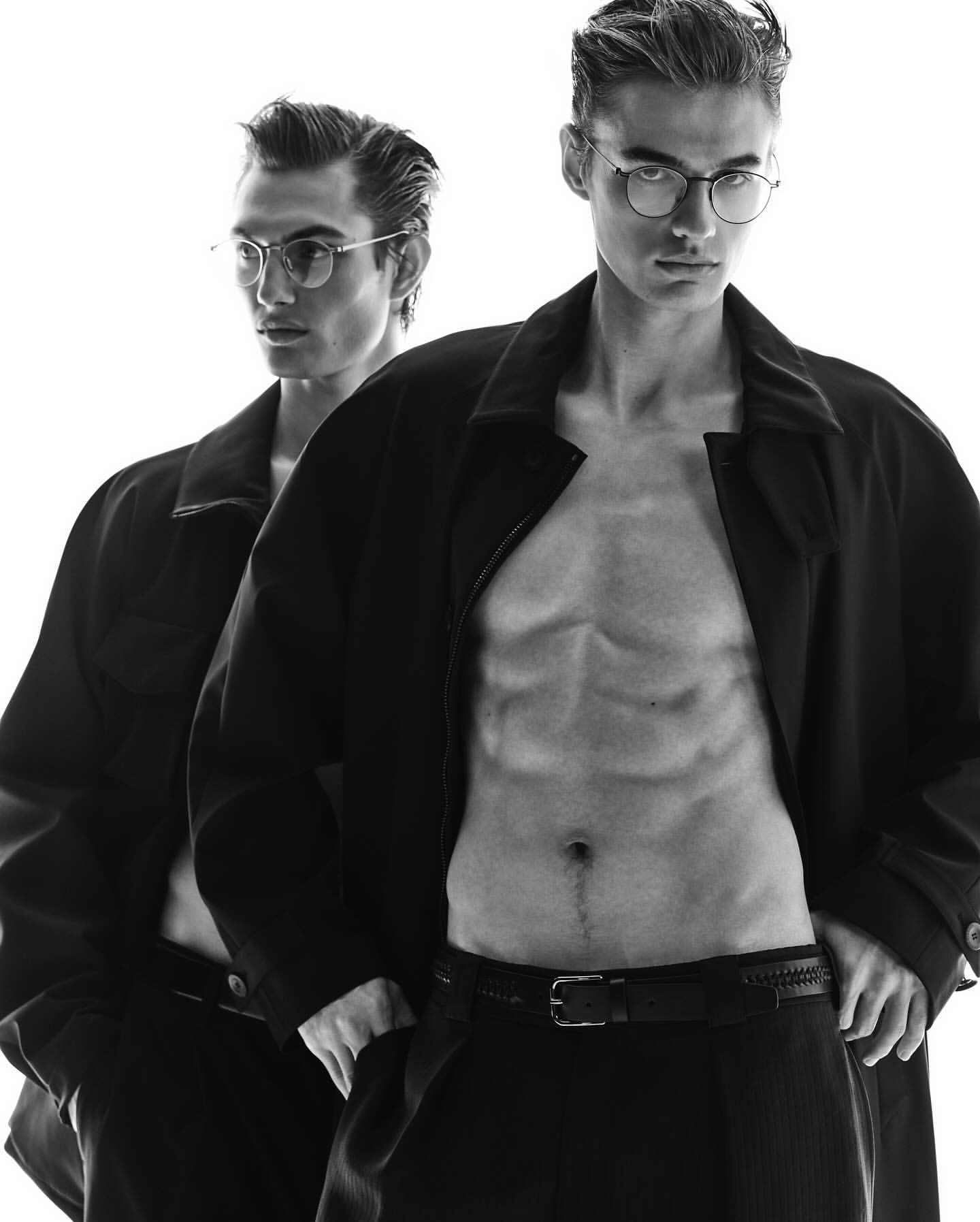 Two adult men pose in a black-and-white studio shot, one shirtless in an unbuttoned coat and dress pants, both wearing glasses.