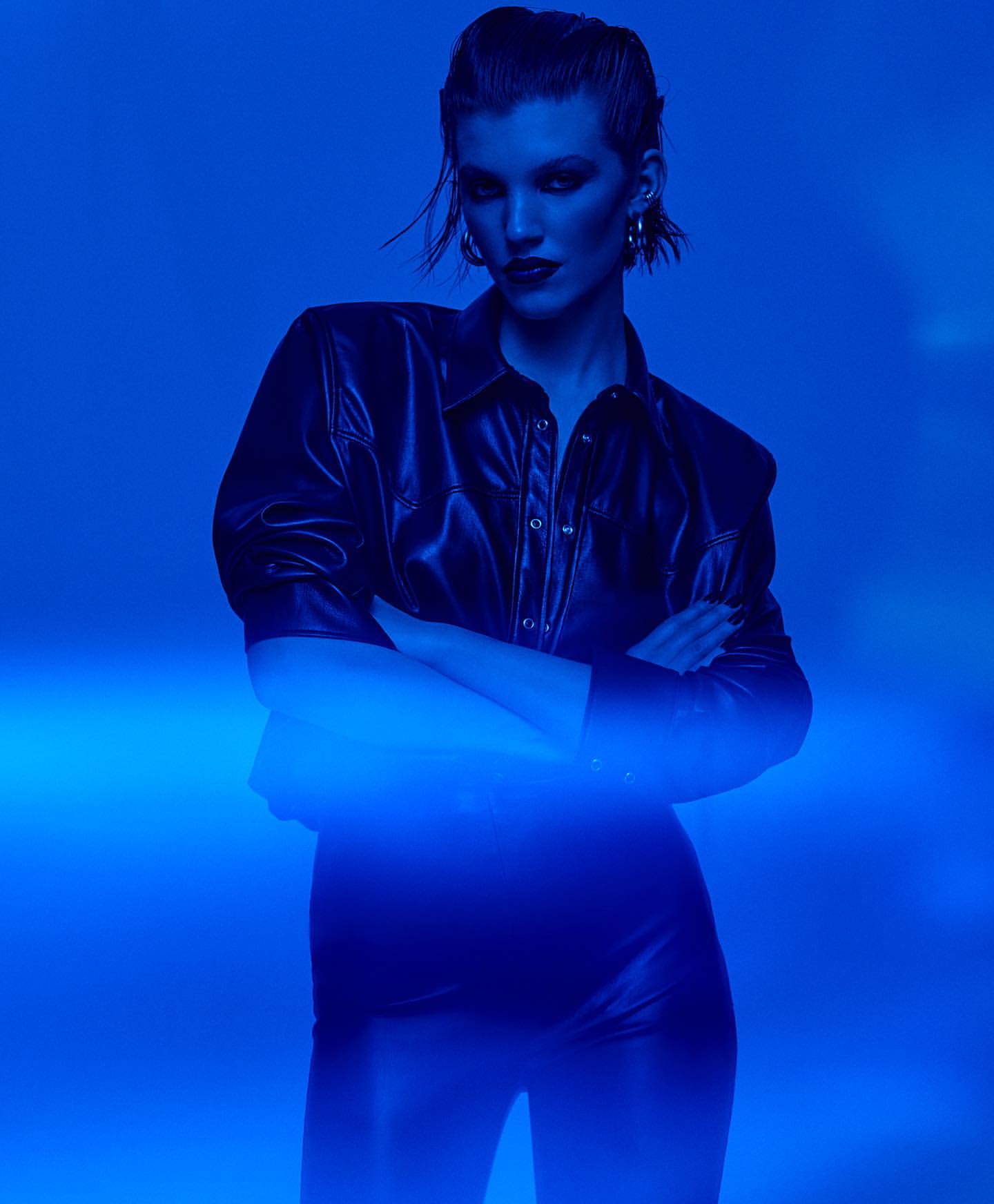 Fashion shot of a woman in a black leather jacket with arms crossed against a bright blue background in a photo studio.