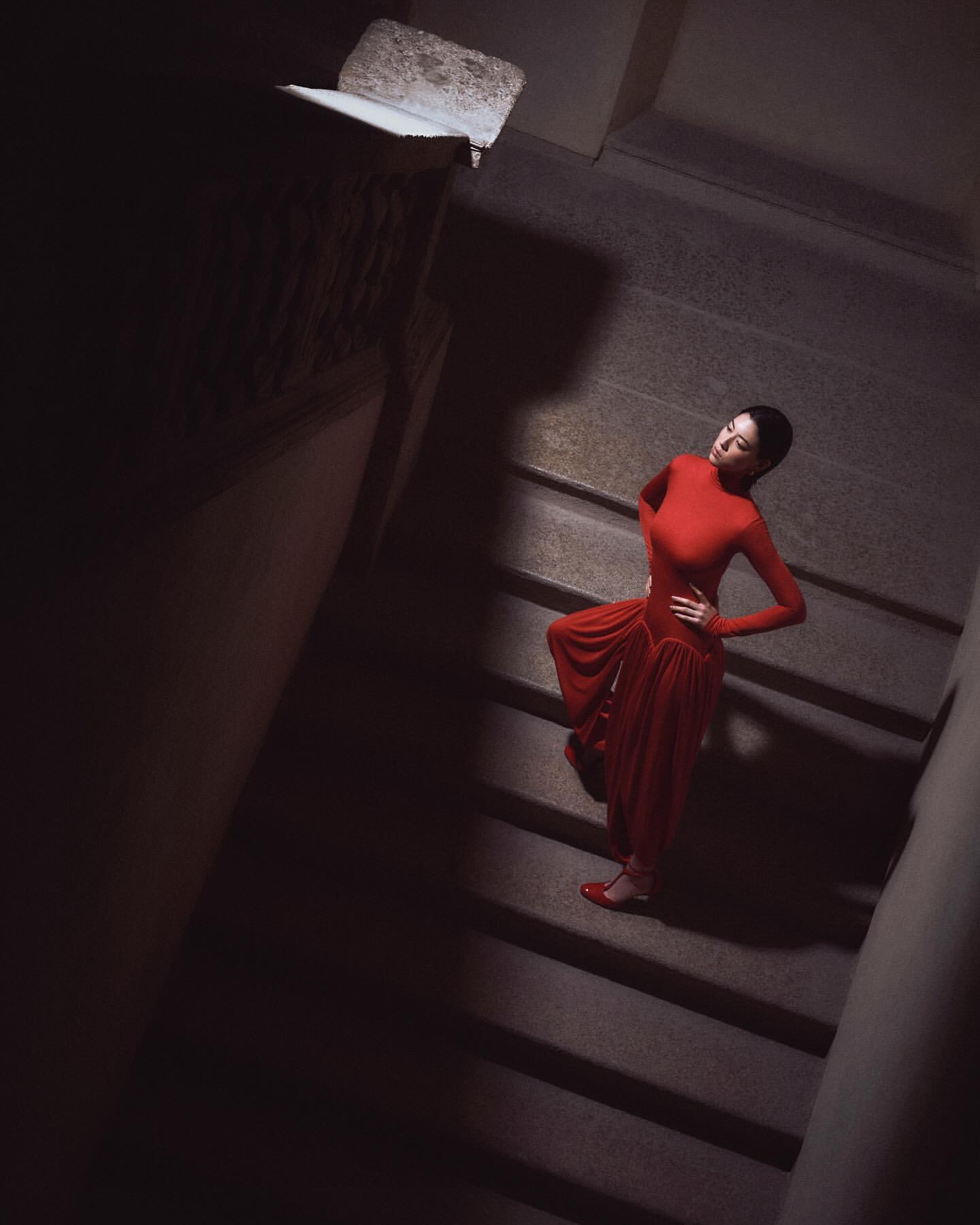 Woman in a red dress poses on a dark stone staircase. She wears matching red shoes and stands with one hand on her hip.