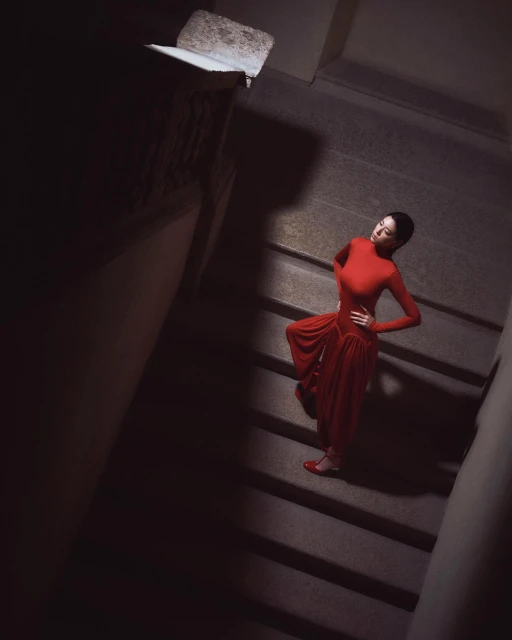 Woman in a red dress poses on a dark stone staircase. She wears matching red shoes and stands with one hand on her hip.