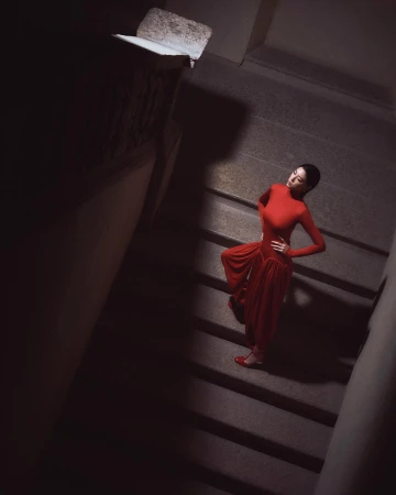 Woman in a long red dress standing on stone stairs under dramatic overhead lighting photographed by Giulio Rustichelli