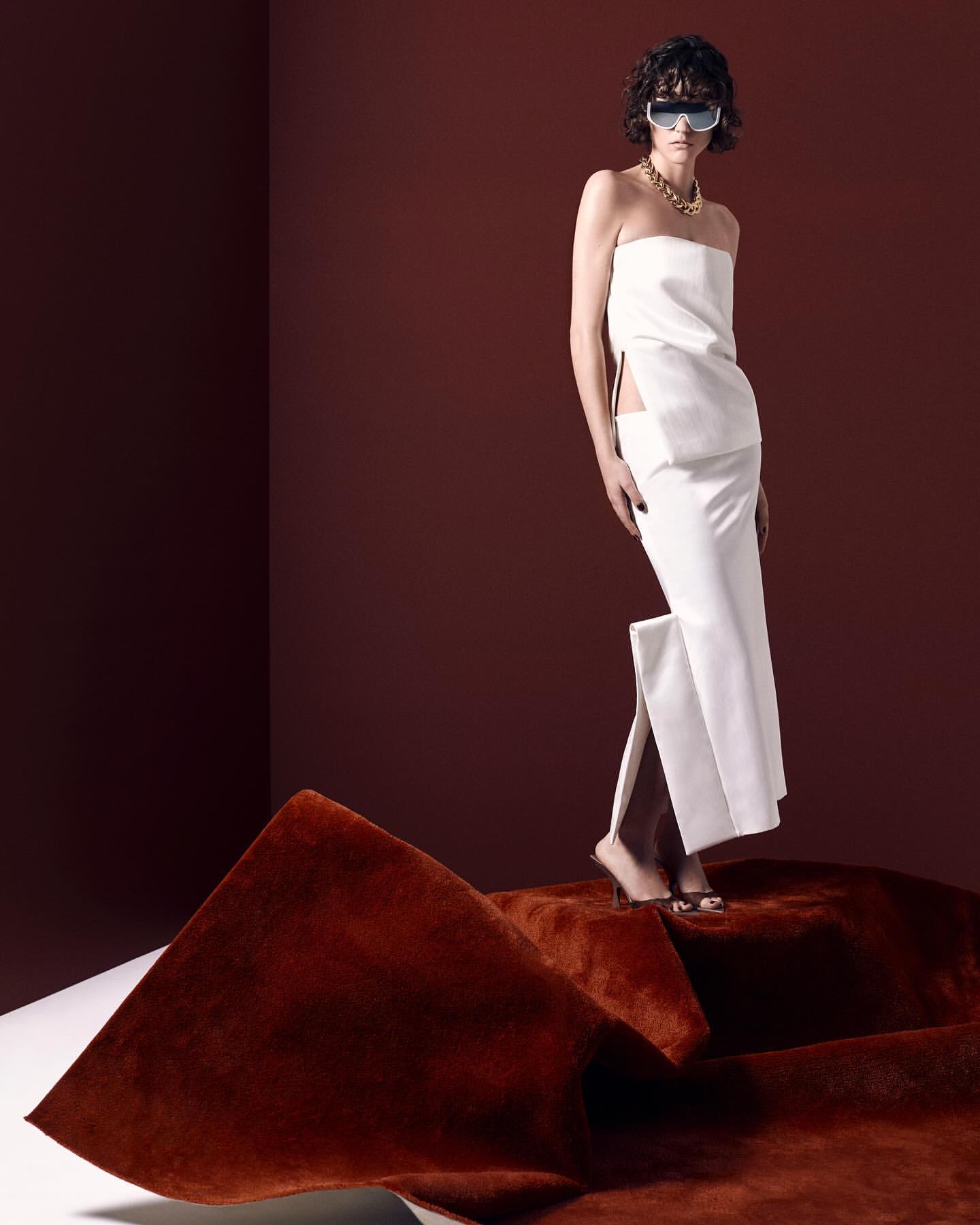 A model poses in a strapless white dress and white sunglasses on a brown surface against a dark red backdrop in a studio fashion shot.