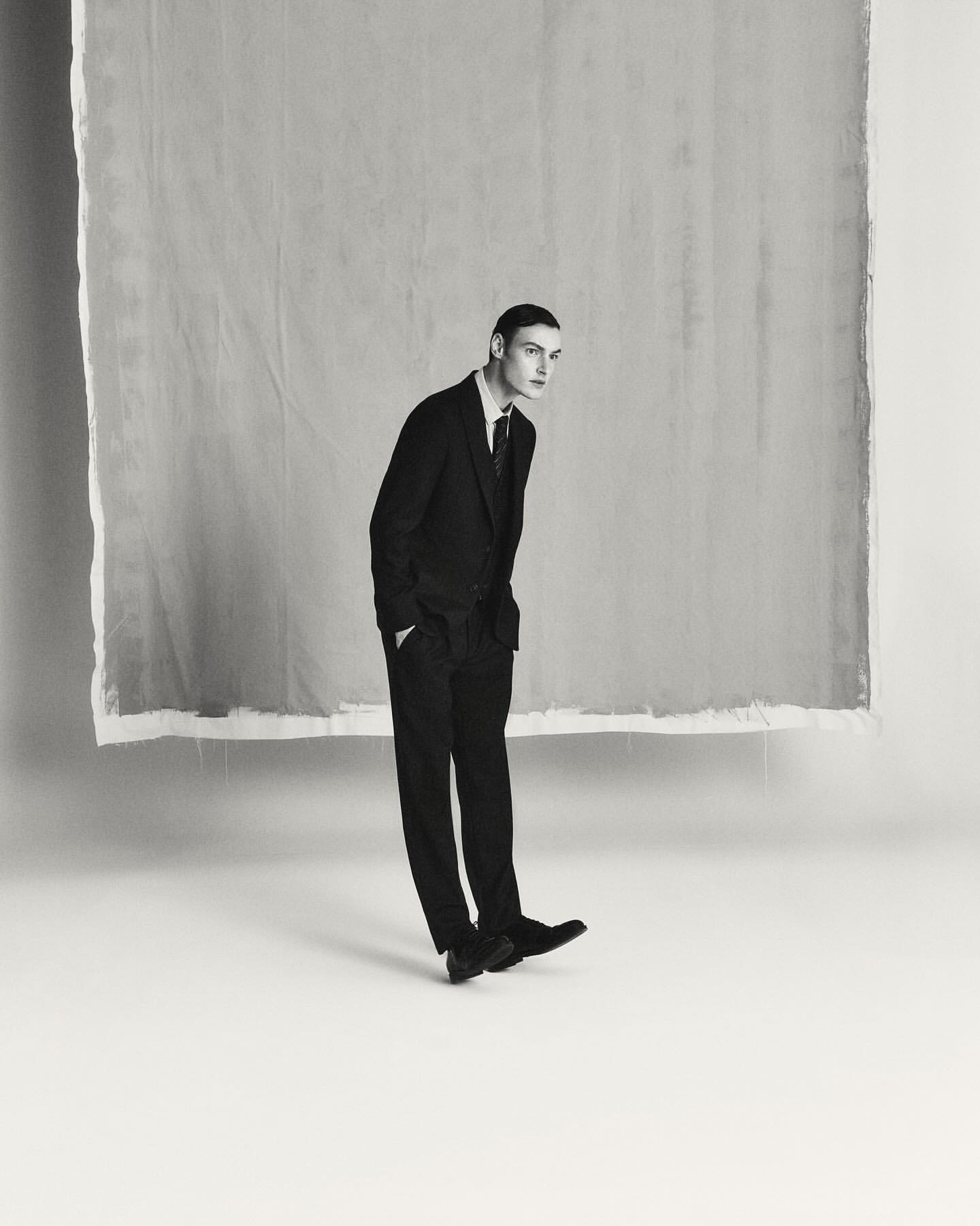 Man wearing a dark suit and tie standing against a light seamless background in a vertical studio portrait