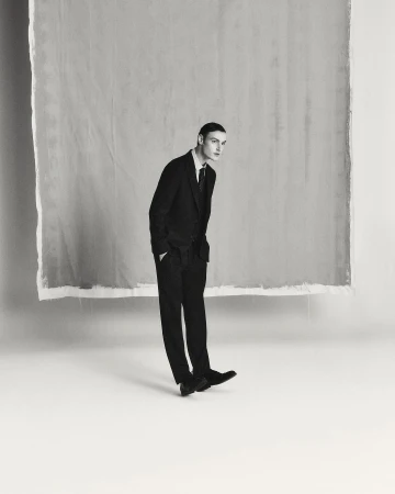 Man wearing a dark suit and tie standing against a light seamless background in a vertical studio portrait